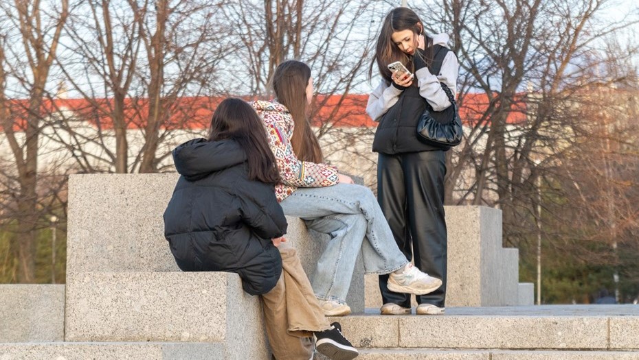 Девушки смотрят в сторону подруги, которая просматривает сообщения на экране смартфона.