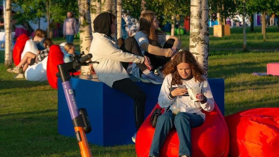 Утомленные зрители и гости фестиваля ВКонтакте VK Fest Санкт-Петербург 2023 отдыхают на пуфиках и надувных креслах близ стендов участников фестиваля, просматривают ленту новостей и вкладывают посты.